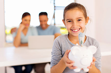 Child smiling and holding piggy bank with money coming out. Parents in the background smiling on computer