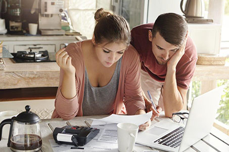 Man and woman at kitchen table looking over papers with coffee, calculator, and laptop.